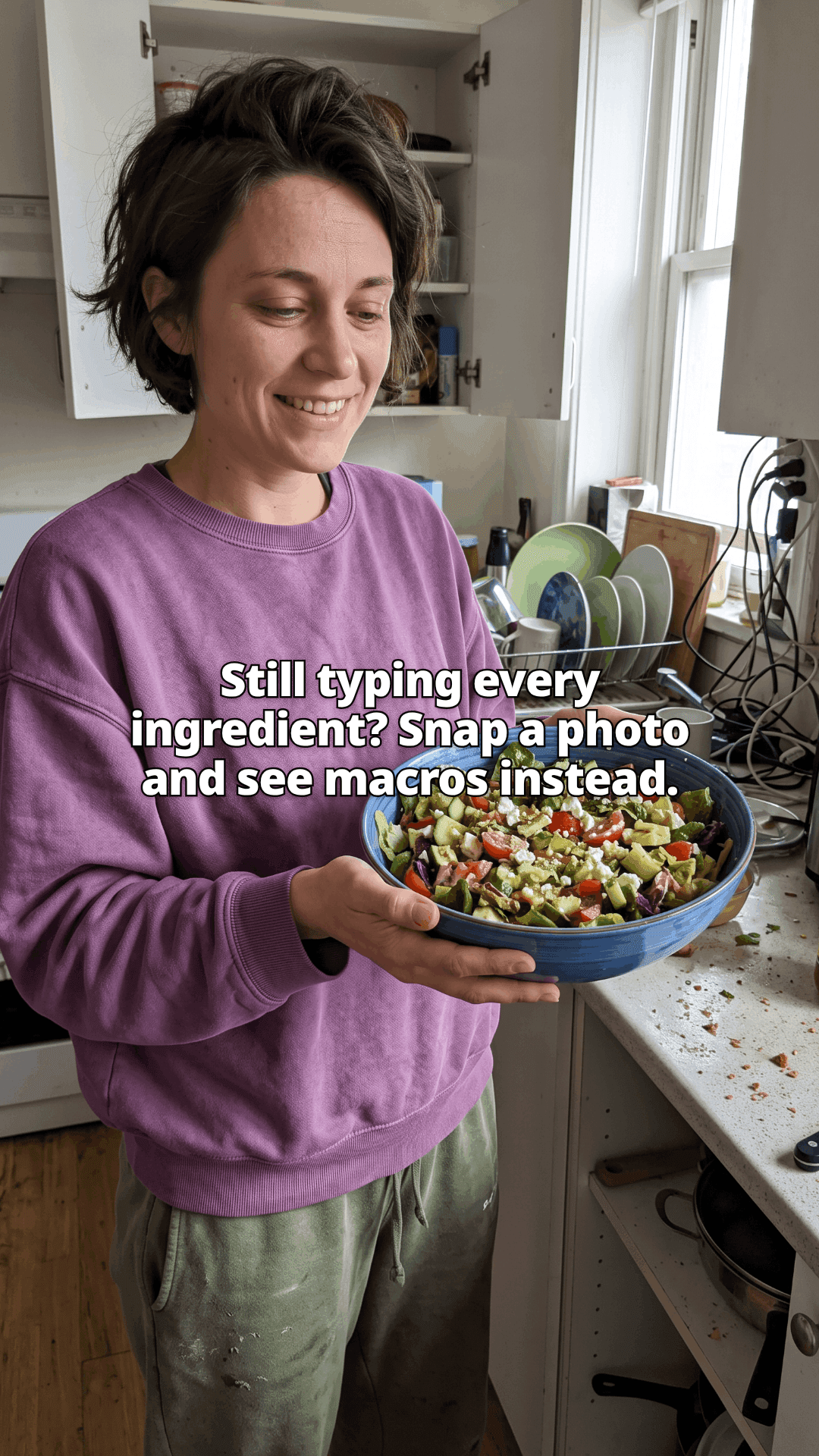 A woman in a purple sweatshirt standing in a cluttered kitchen, smiling down at the large blue bowl of chopped salad she is holding, with the opening line of the post across the photo.