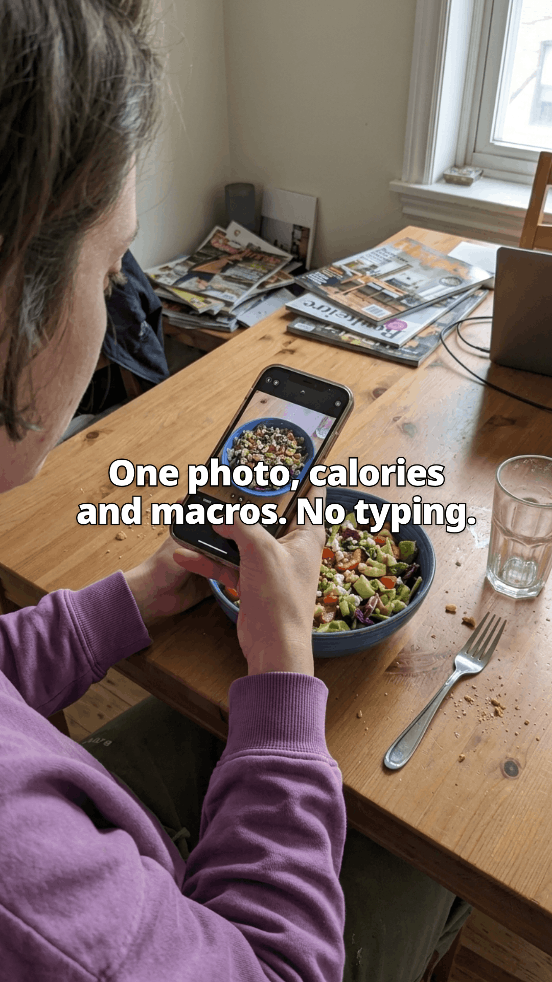Seen over a woman's shoulder at a wooden table, she holds her phone above a bowl of salad to photograph it, a fork and a glass of water beside it, with a line of the post across the photo.