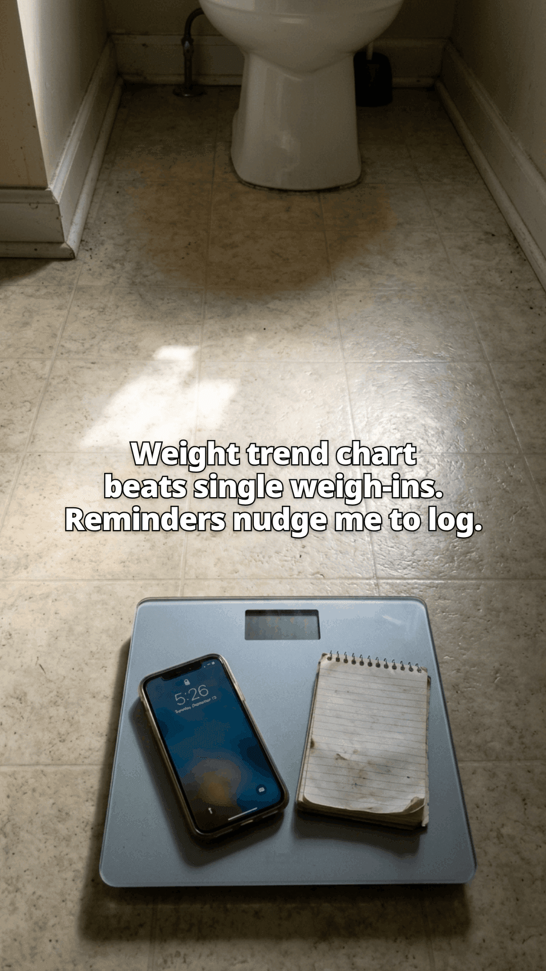 A bathroom scale on a tiled floor with a phone and a spiral notepad resting on it, in a patch of low sunlight, with a line of the post across the photo.